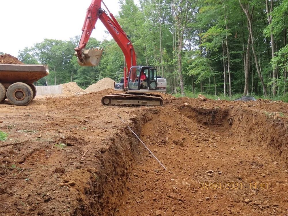Excavator digging a trench in brown dirt, a dump truck nearby, forest in the background.