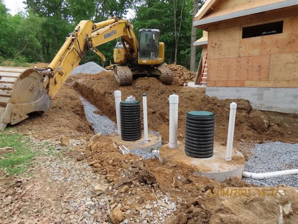 Excavator digging near septic tank installation. Construction site near a building, soil, gravel, and pipes are visible.