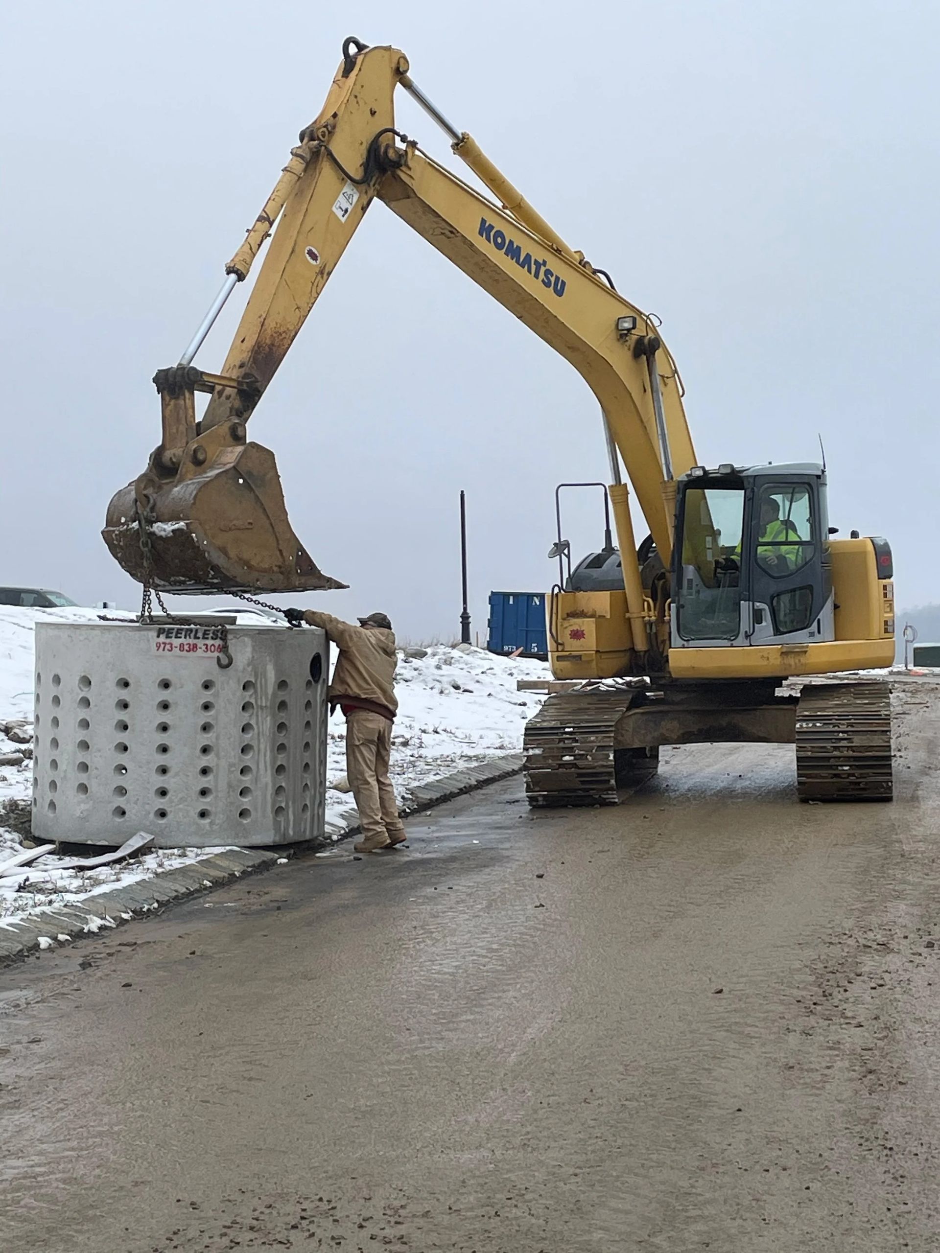 Yellow excavator lifting a concrete box; worker guides it on a paved road. Snowy setting.