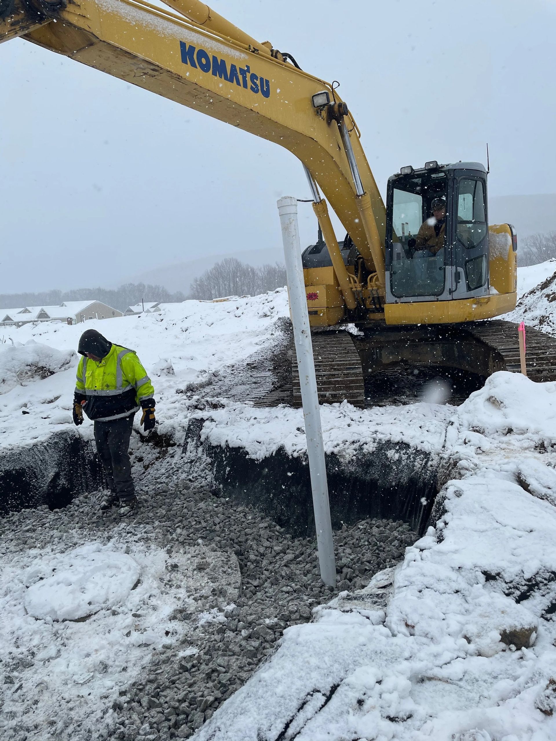 Yellow excavator digging a trench in snowy conditions; a worker in a safety vest observes.