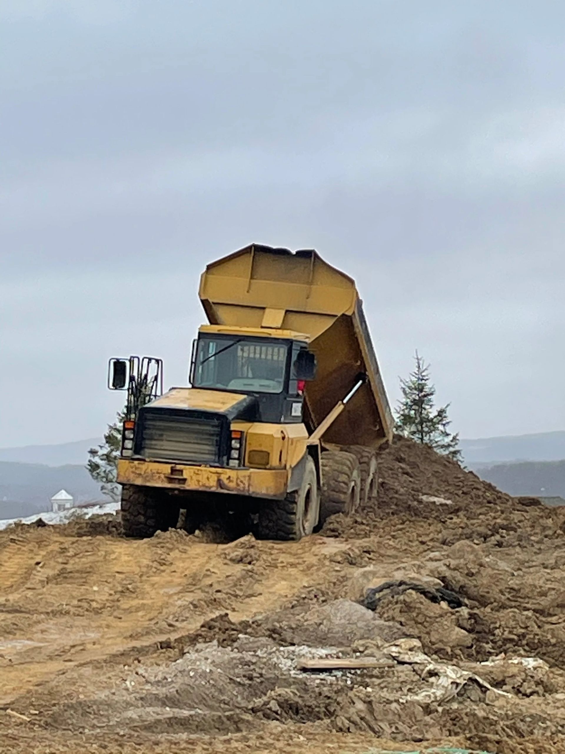 Yellow dump truck unloading dirt on a construction site under a cloudy sky.