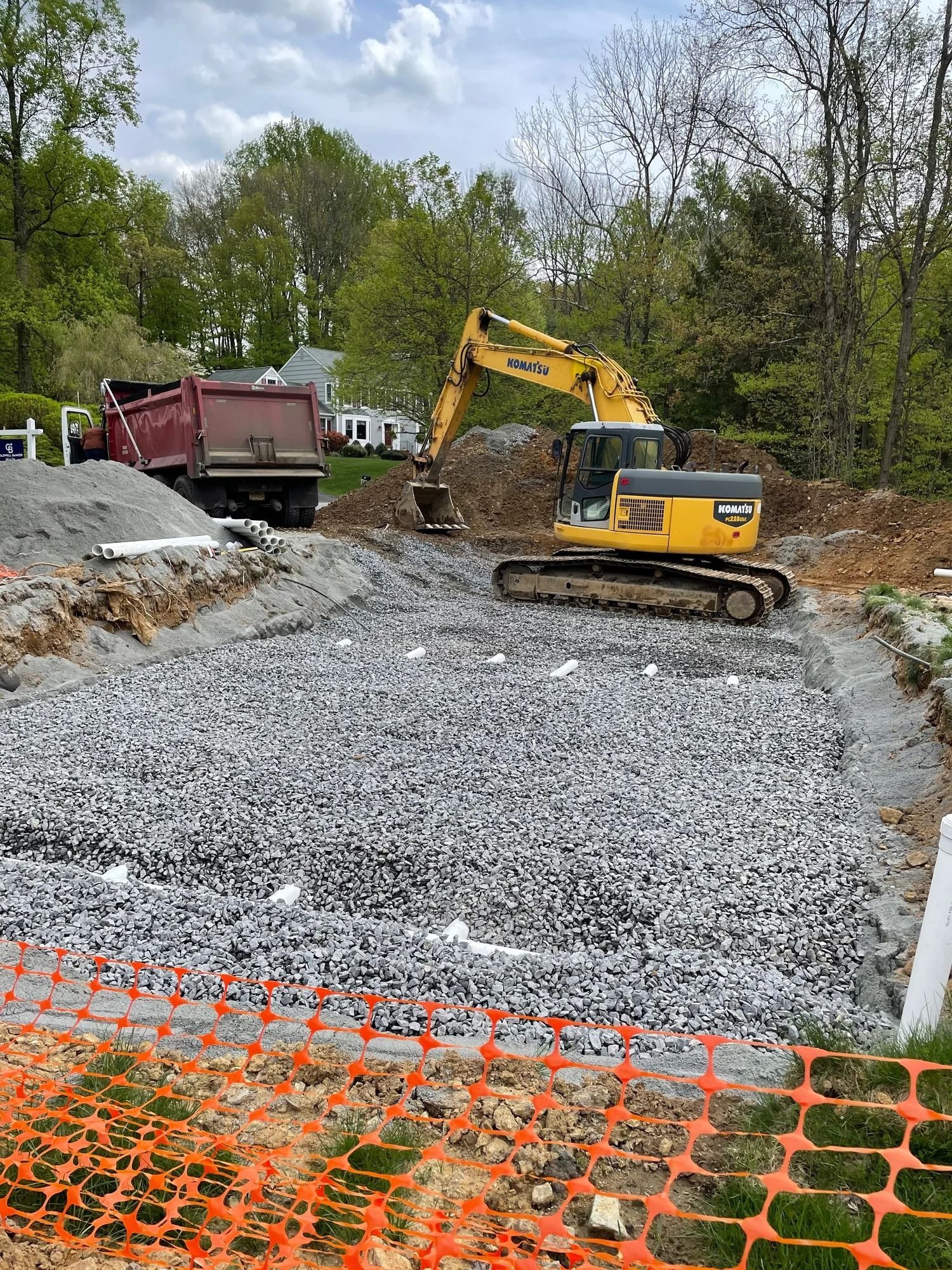 Construction site with an excavator, gravel bed, and dump truck; orange safety fence.