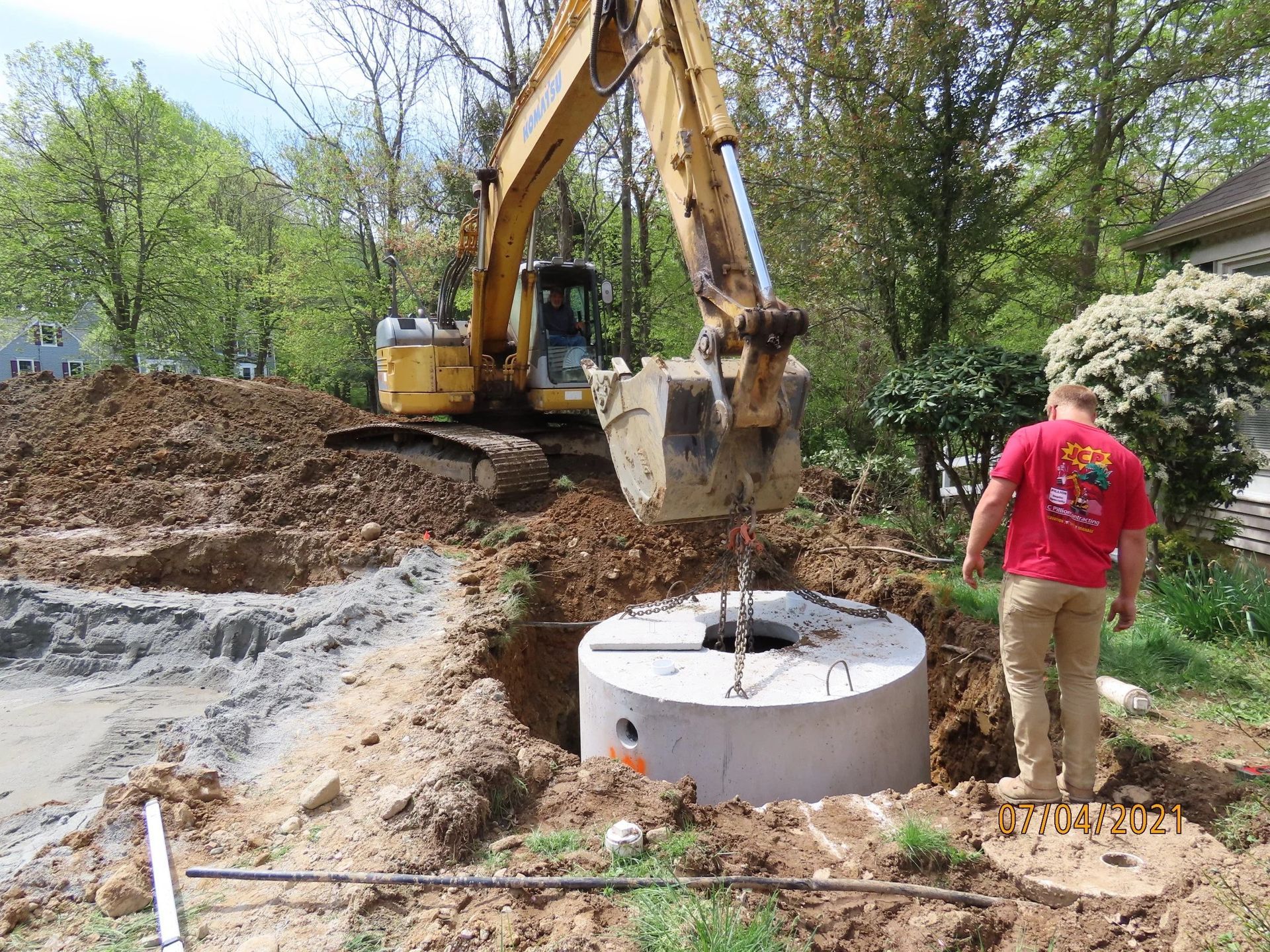 An excavator lowering a concrete septic tank into a hole with a worker nearby on a residential construction site.