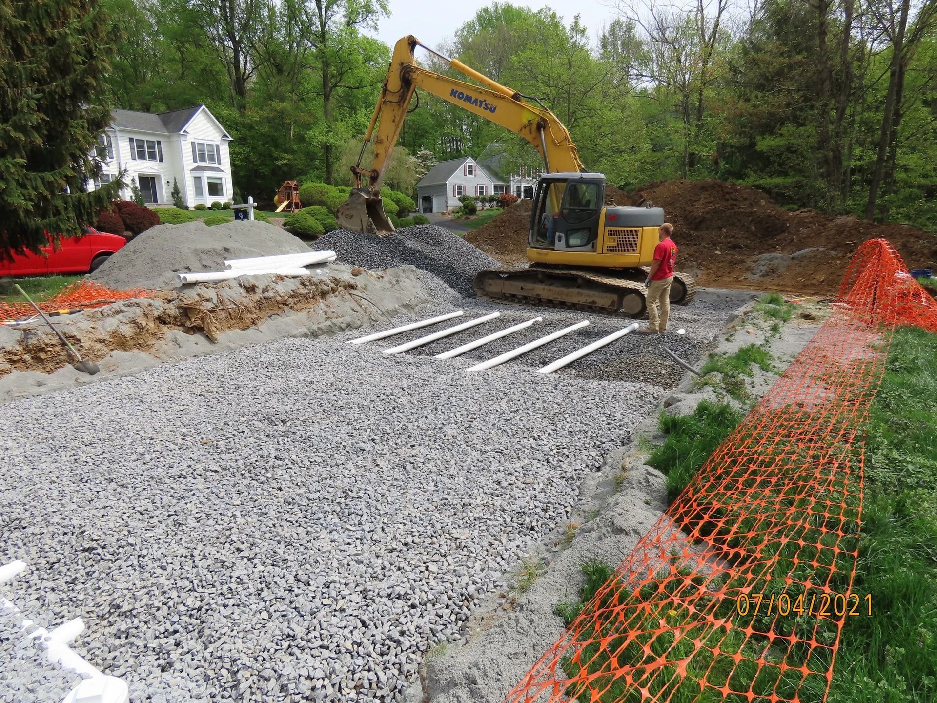 Excavator working on a gravel-filled drainage area with pipes and orange safety fencing in a residential yard.