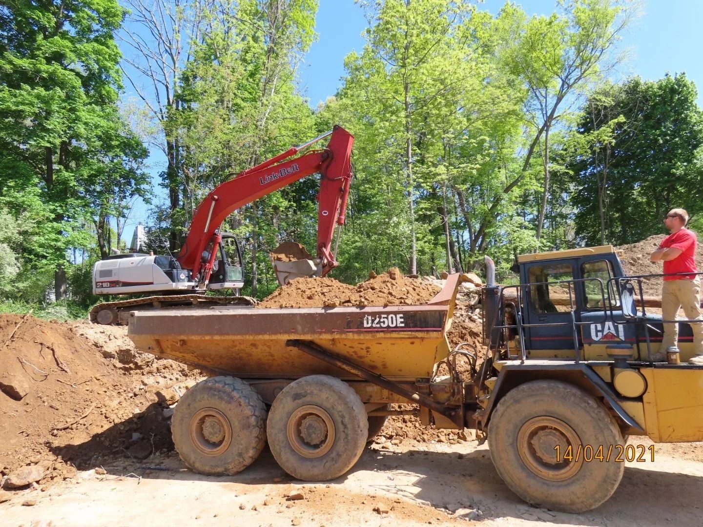 An excavator loads dirt into a yellow dump truck on a sunny day; a man stands on the truck.