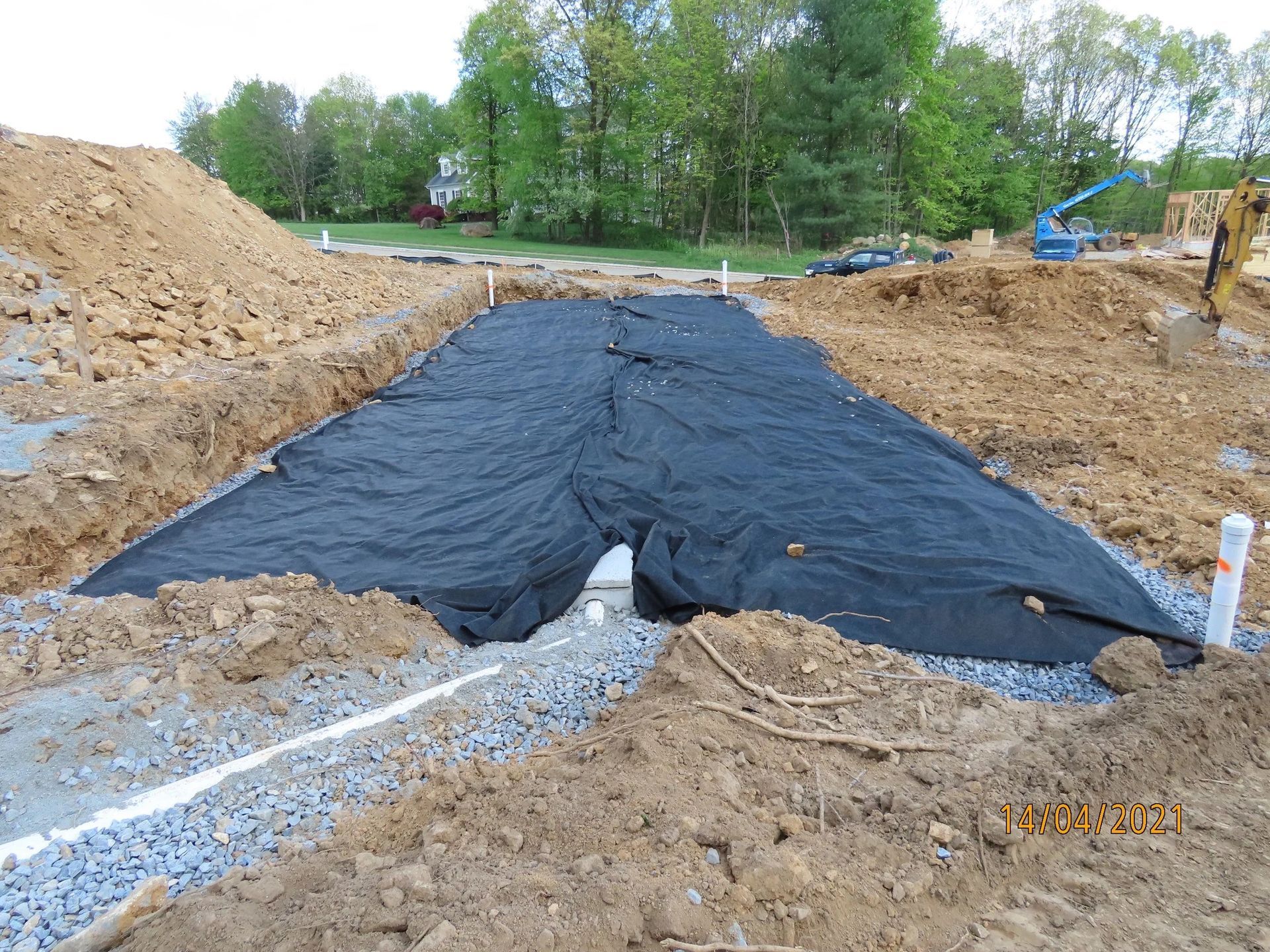 Construction site, dark fabric covering gravel-filled trench, surrounded by dirt and pipes, trees in the background.