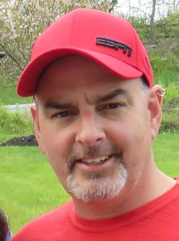 Man wearing red ESPN hat and shirt smiles outdoors.