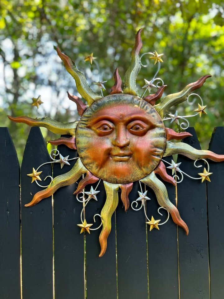 Metal sun face decoration with stars on a black fence. The sun has a smiling face and a rusty orange and yellow color.