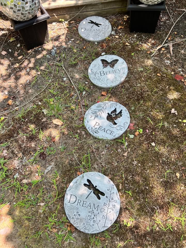 Stone stepping stones in a garden path, each inscribed with inspirational words and a nature-themed silhouette: dragonfly, butterfly, or bird.