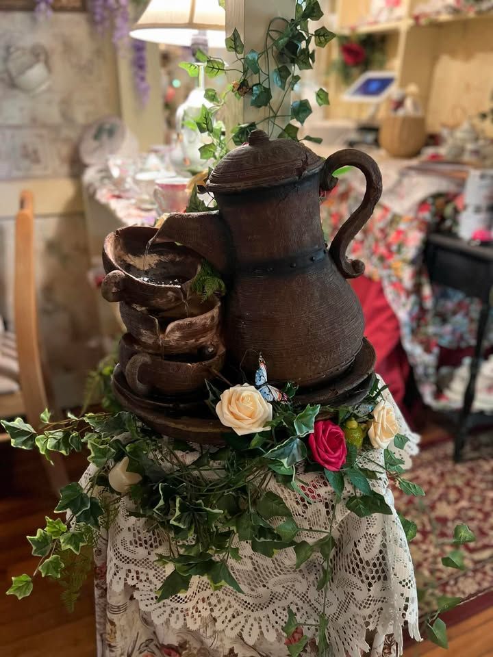 Water fountain shaped like a teapot pouring into stacked cups, surrounded by flowers and ivy.