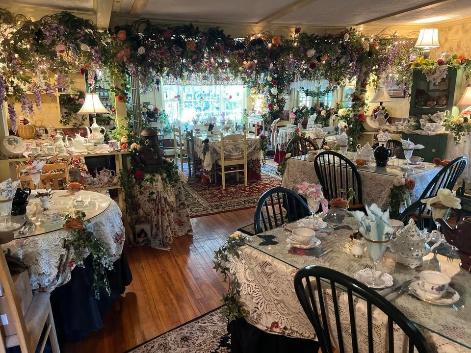 A floral-decorated tea room with tables set for diners. Wooden floors, flowers, and hanging vines create an ornate interior.