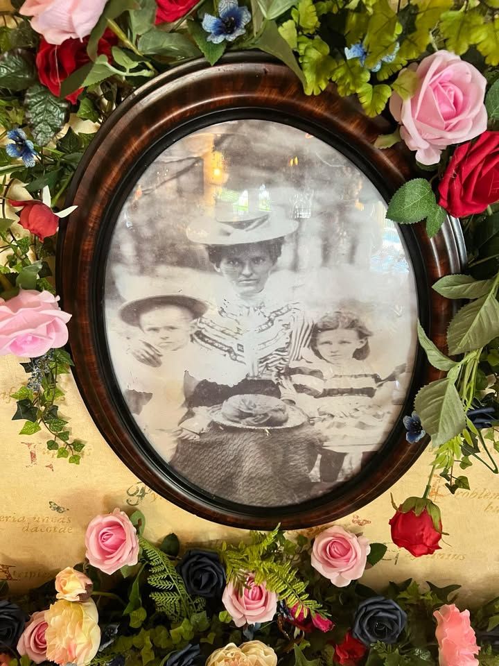 Black and white oval framed photo of a woman and two children, surrounded by a floral wreath of pink, red and blue roses.