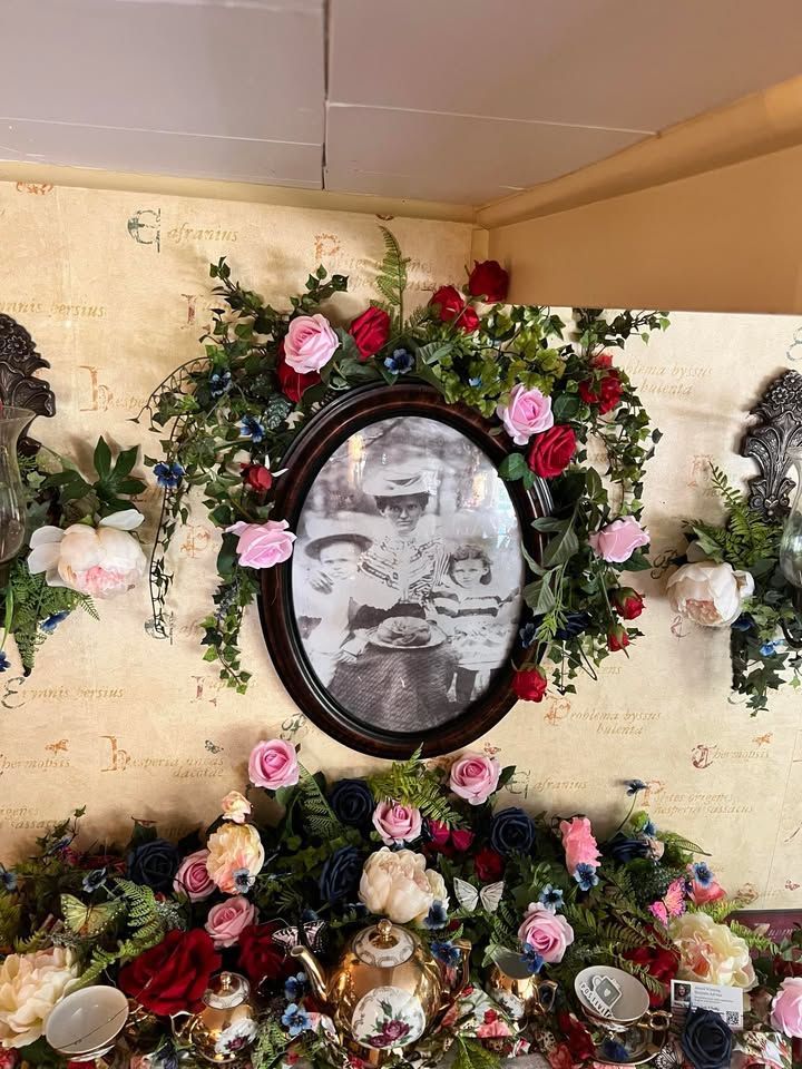 Oval framed black and white photo surrounded by flowers and greenery on a textured yellow wall.
