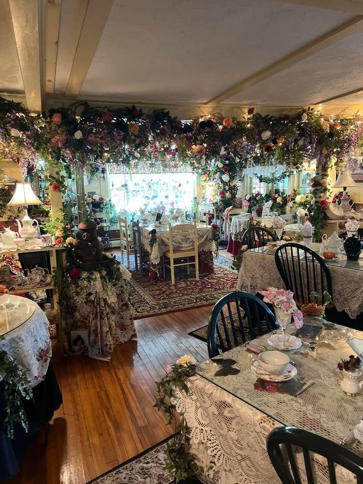 A room decorated with flowers and lace-covered tables, set for a tea party. The ceiling is adorned with more flowers and lights.