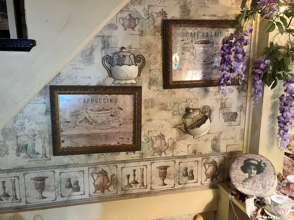 A dining room lavishly decorated with flowers. Tables are set with delicate china and lace, with floral arrangements above and around the window and walls. Wall decorated with framed coffee-themed art and wallpaper with purple floral accent.