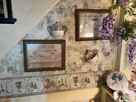 A dining room lavishly decorated with flowers. Tables are set with delicate china and lace, with floral arrangements above and around the window and walls. Wall decorated with framed coffee-themed art and wallpaper with purple floral accent.
