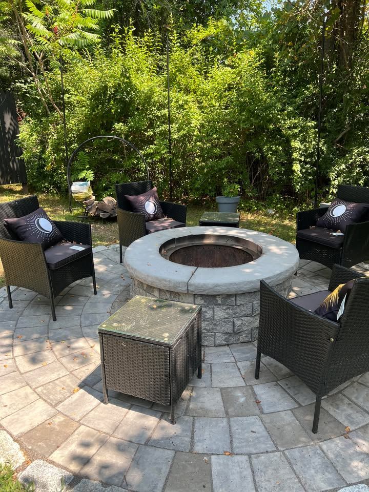 A circular patio with a fire pit surrounded by four wicker chairs with pillows and a side table. The setting is a sunny backyard.