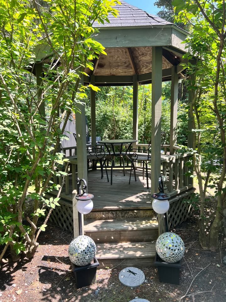 A wooden gazebo with steps, surrounded by trees. Inside are chairs and a table.  Decorative globe lights and stepping stones lead to the gazebo.
