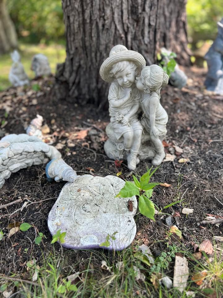 Two small statues of children beside a tree in a garden. One statue wears a hat, the other whispers.