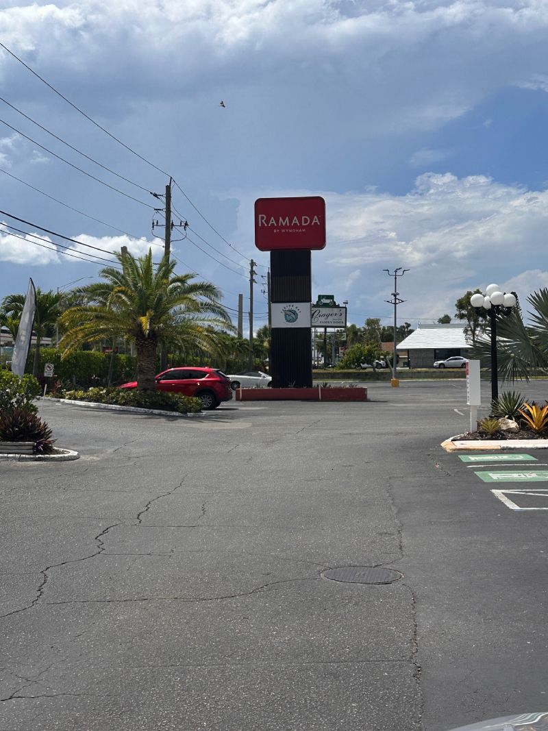 A red and black roadside sign for a restaurant, with a red car parked nearby on a sunny day.
