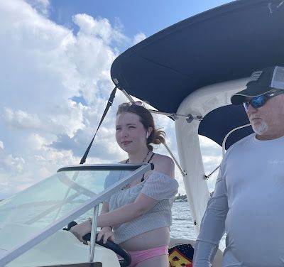 Woman steers a boat on a sunny day; older man looks on. Sky and water visible; both are outdoors.