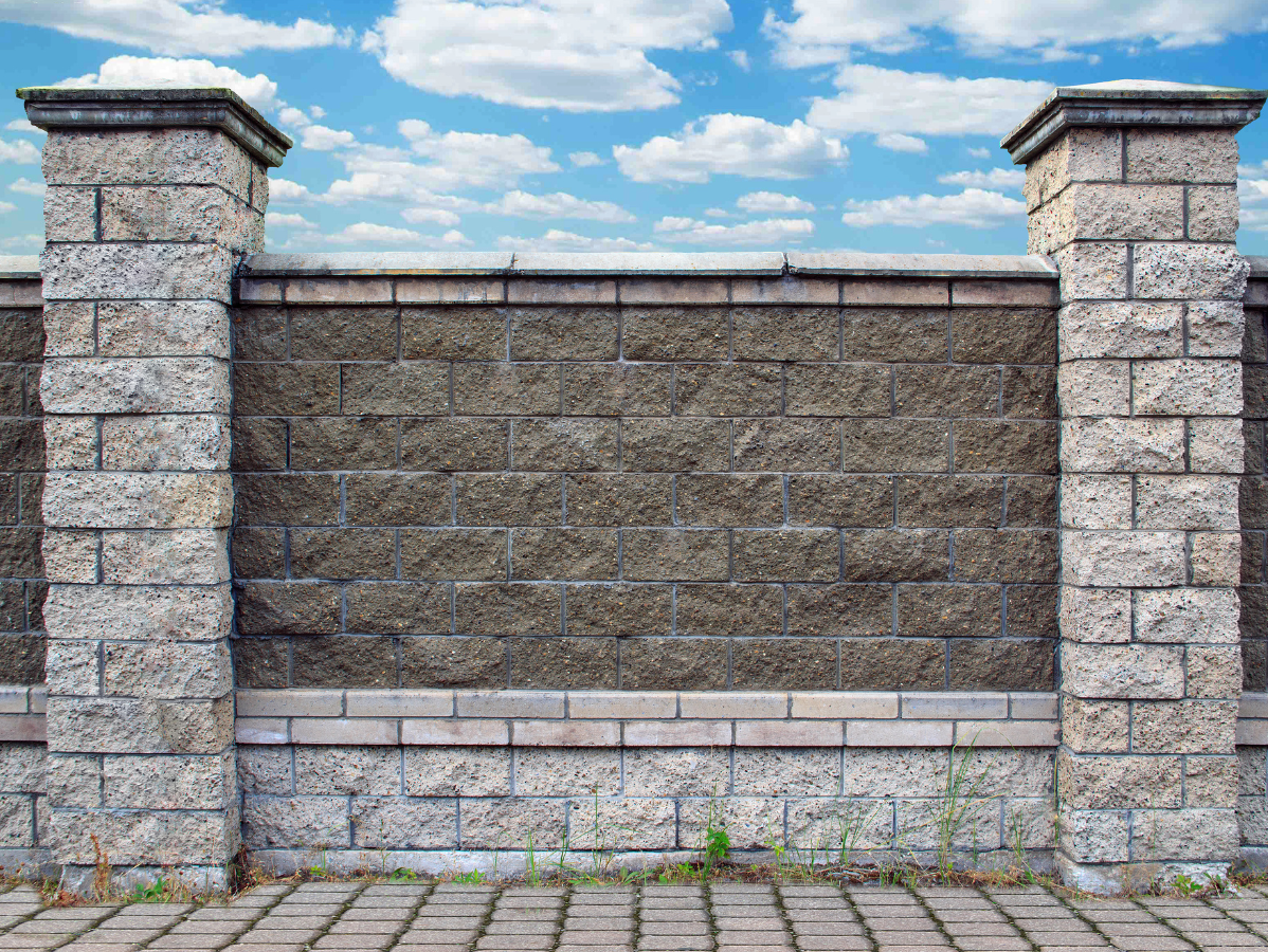 Stone brick fence with decorative pillars against a blue sky with clouds.