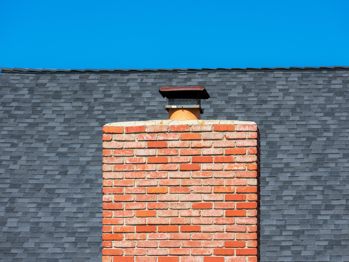 Brick chimney on a gray-shingled roof against a bright blue sky.