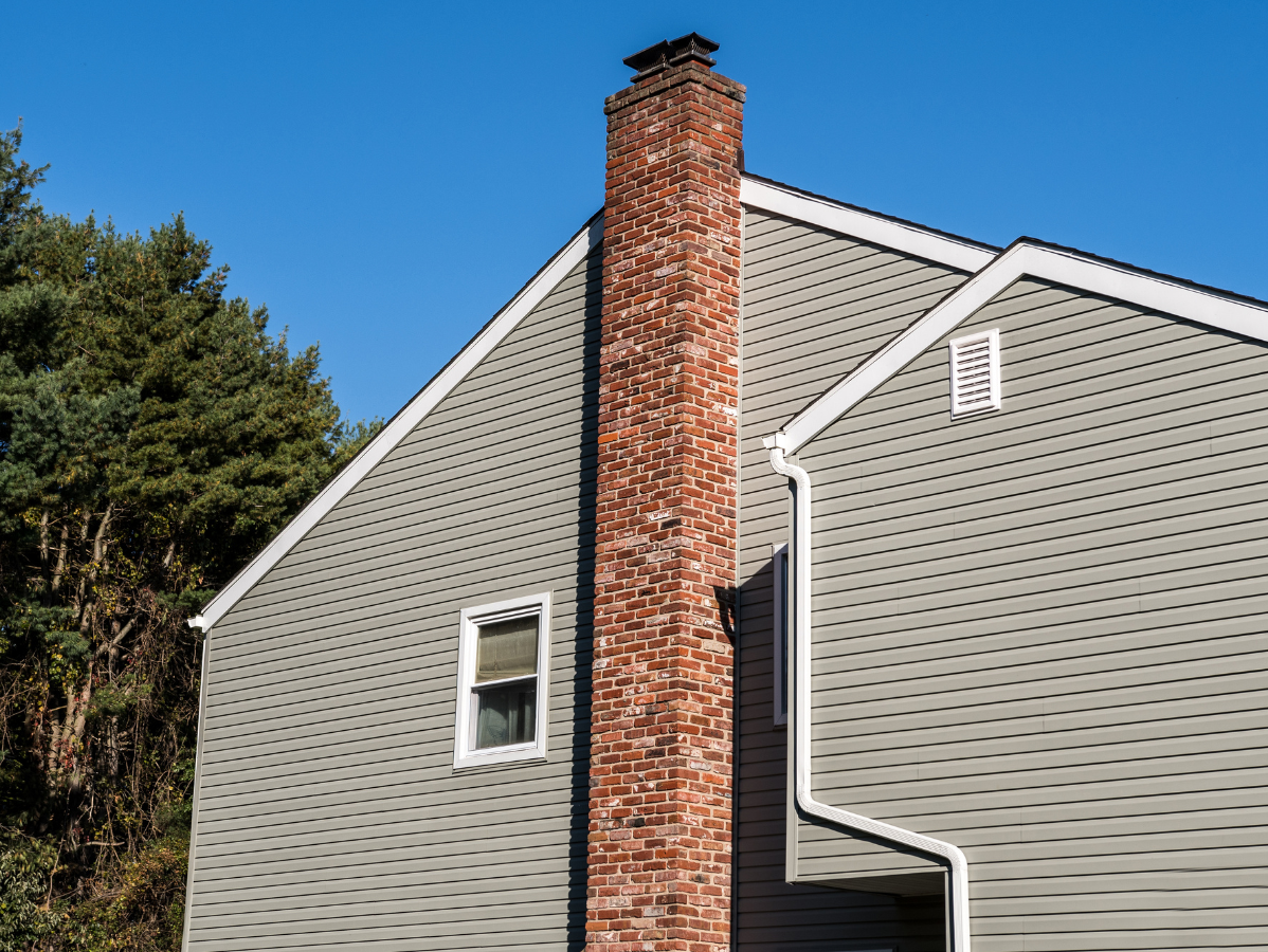 Brick chimney on a gray-sided house, with white trim and a blue sky background.