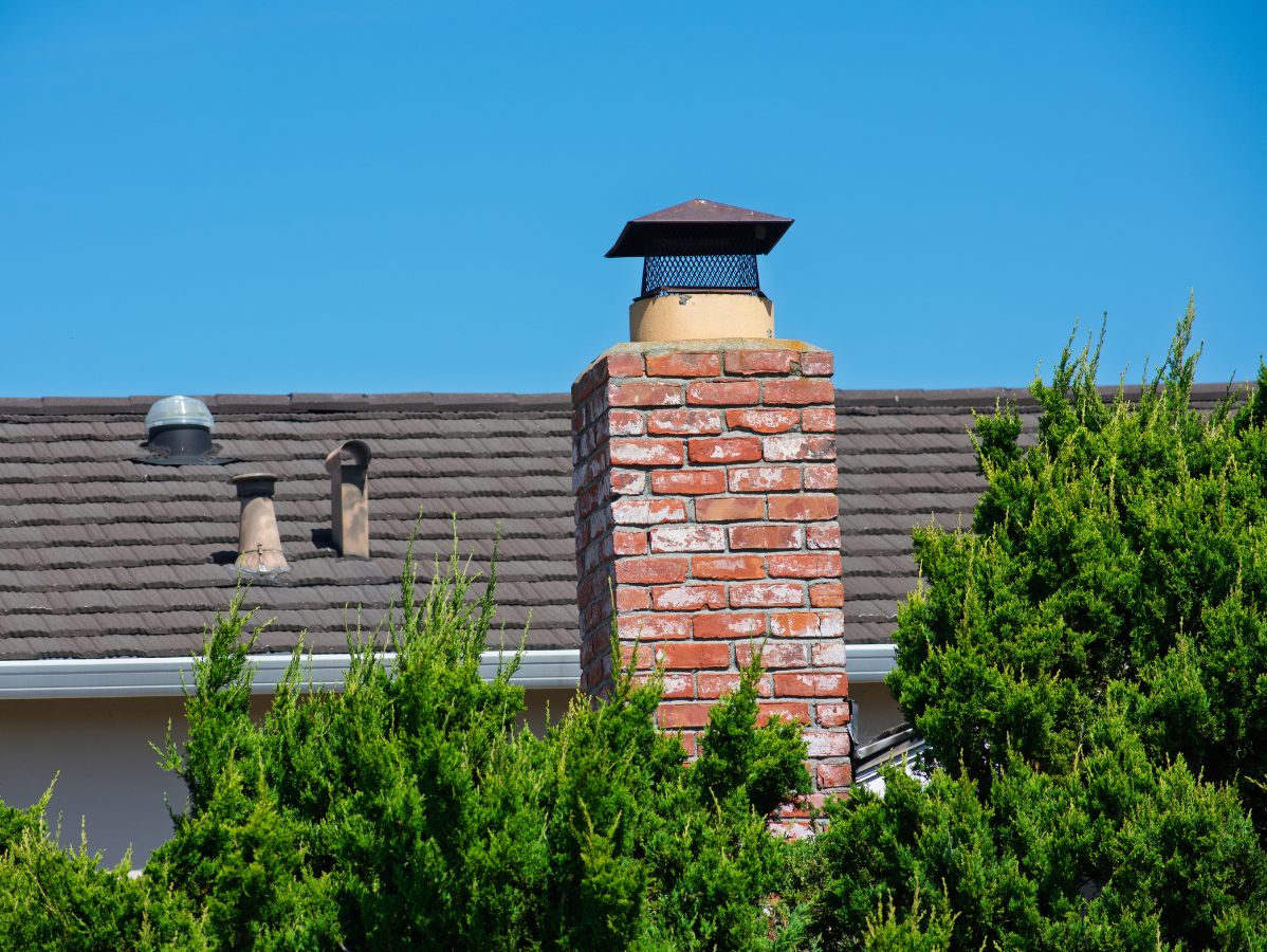 Brick chimney with metal cap on a roof, surrounded by green bushes, against a clear blue sky.