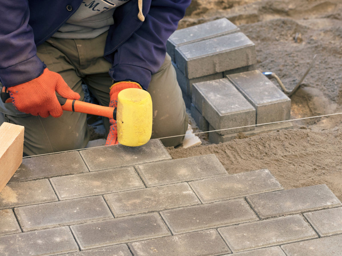 Person in orange gloves laying bricks with a mallet on a sandy surface.