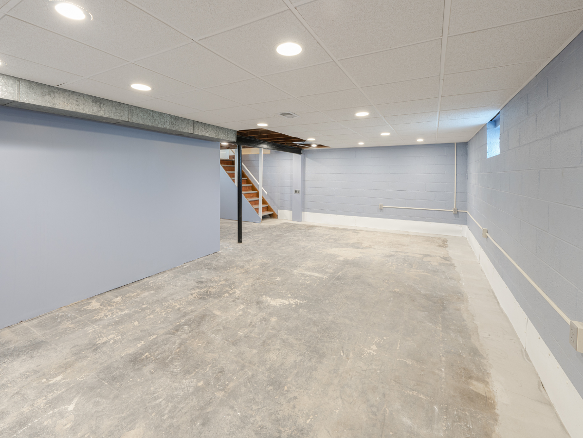 Empty, light blue basement with concrete floor; stairs in the back, recessed lighting, and a small window.