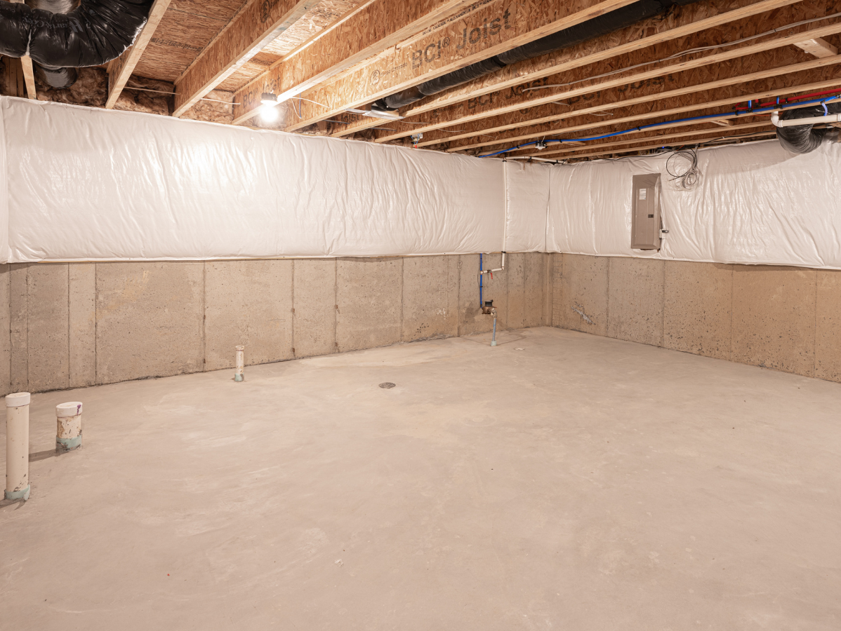 Unfinished basement with concrete walls, wooden ceiling beams, and plumbing.