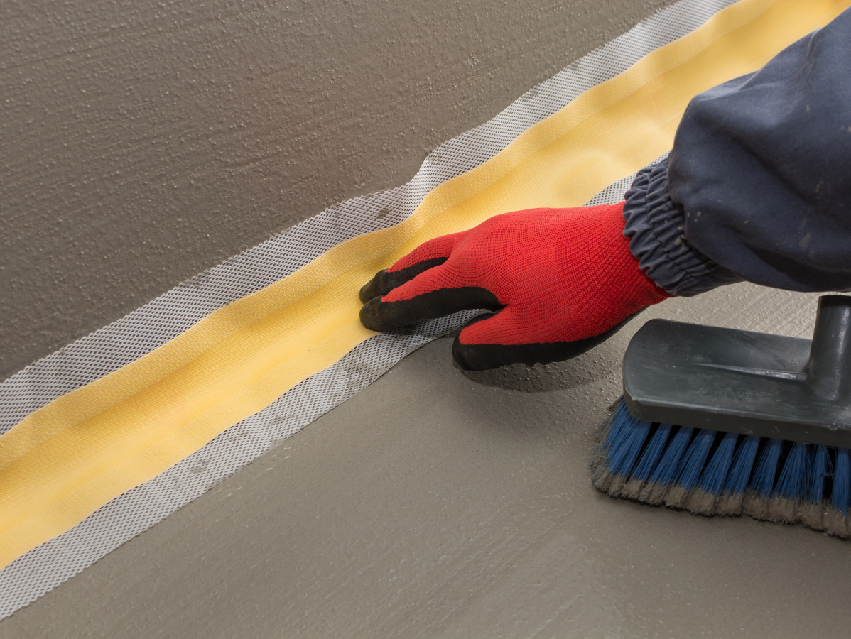A gloved hand smoothing tape with a brush on a concrete surface for sealing.