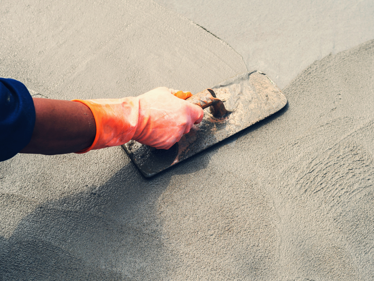 Person wearing orange gloves using a trowel to smooth cement on a light gray surface.