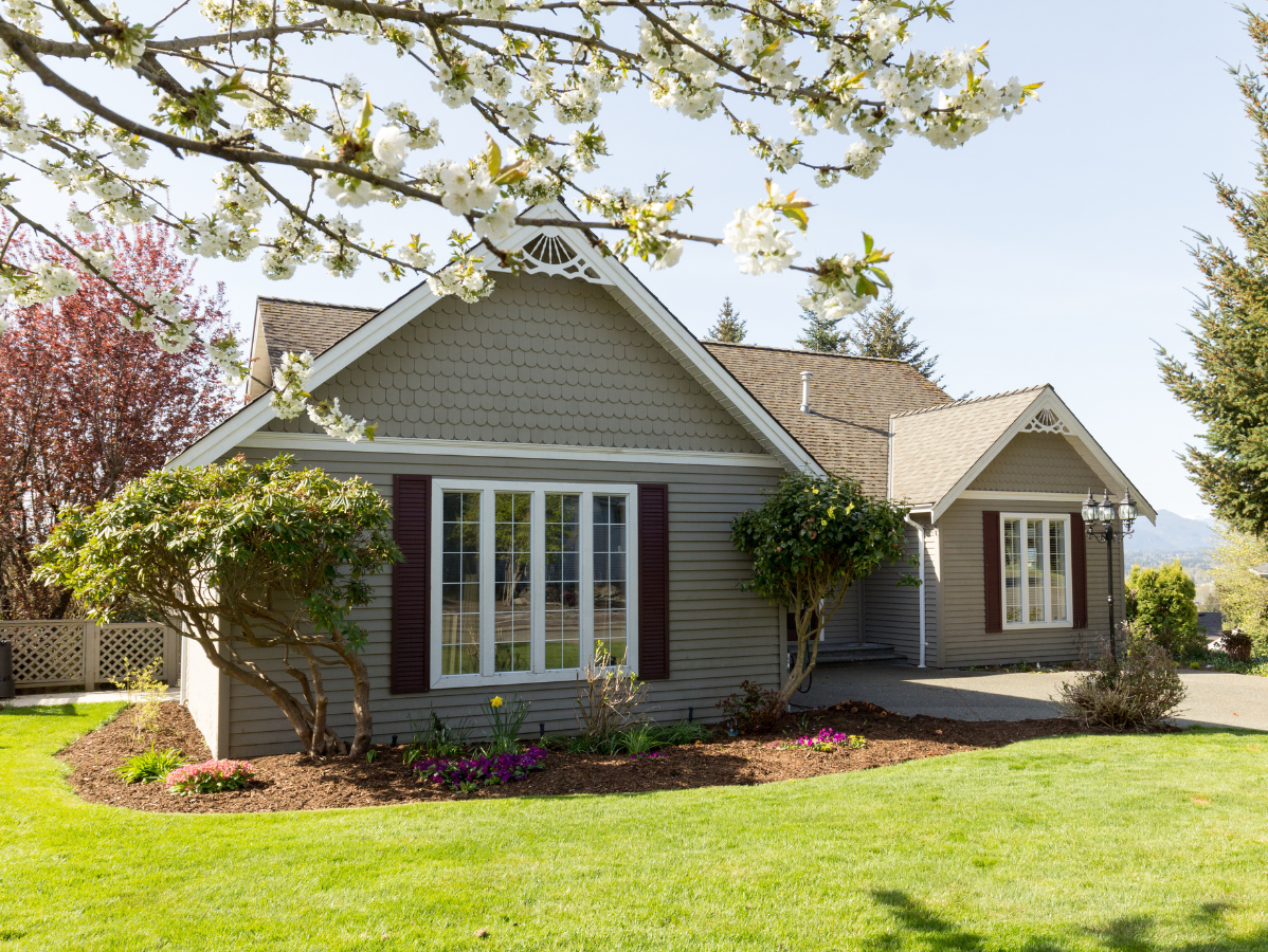 Cozy gray house with white trim, dark red shutters, and a green lawn, under a blossoming tree.