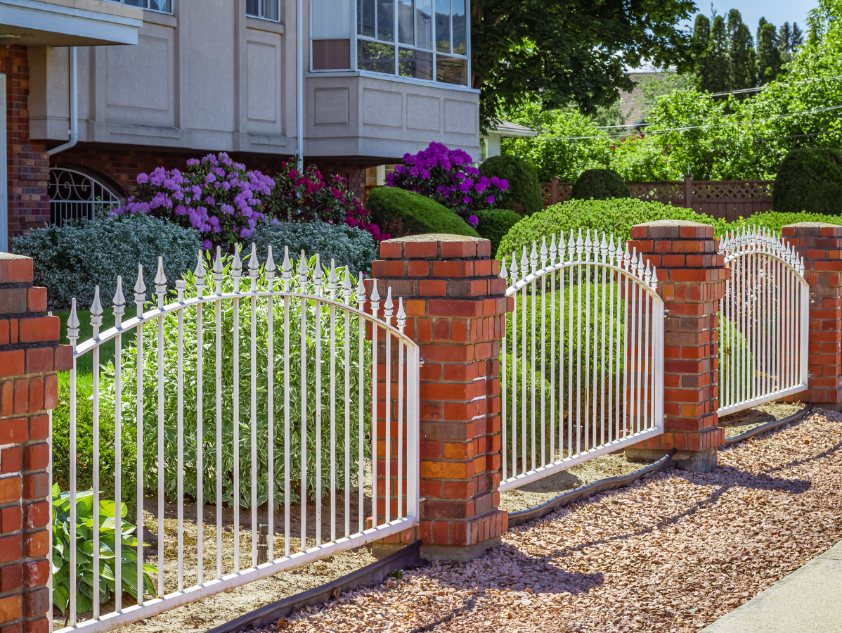 White metal fence with red brick pillars in front of a house with a garden of bushes and flowers.