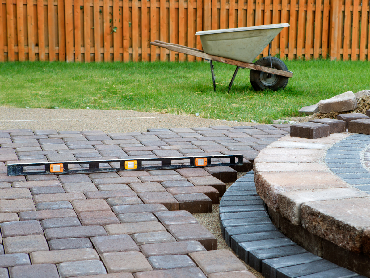 Construction of a brick patio and fire pit in a backyard, with a level and wheelbarrow visible.