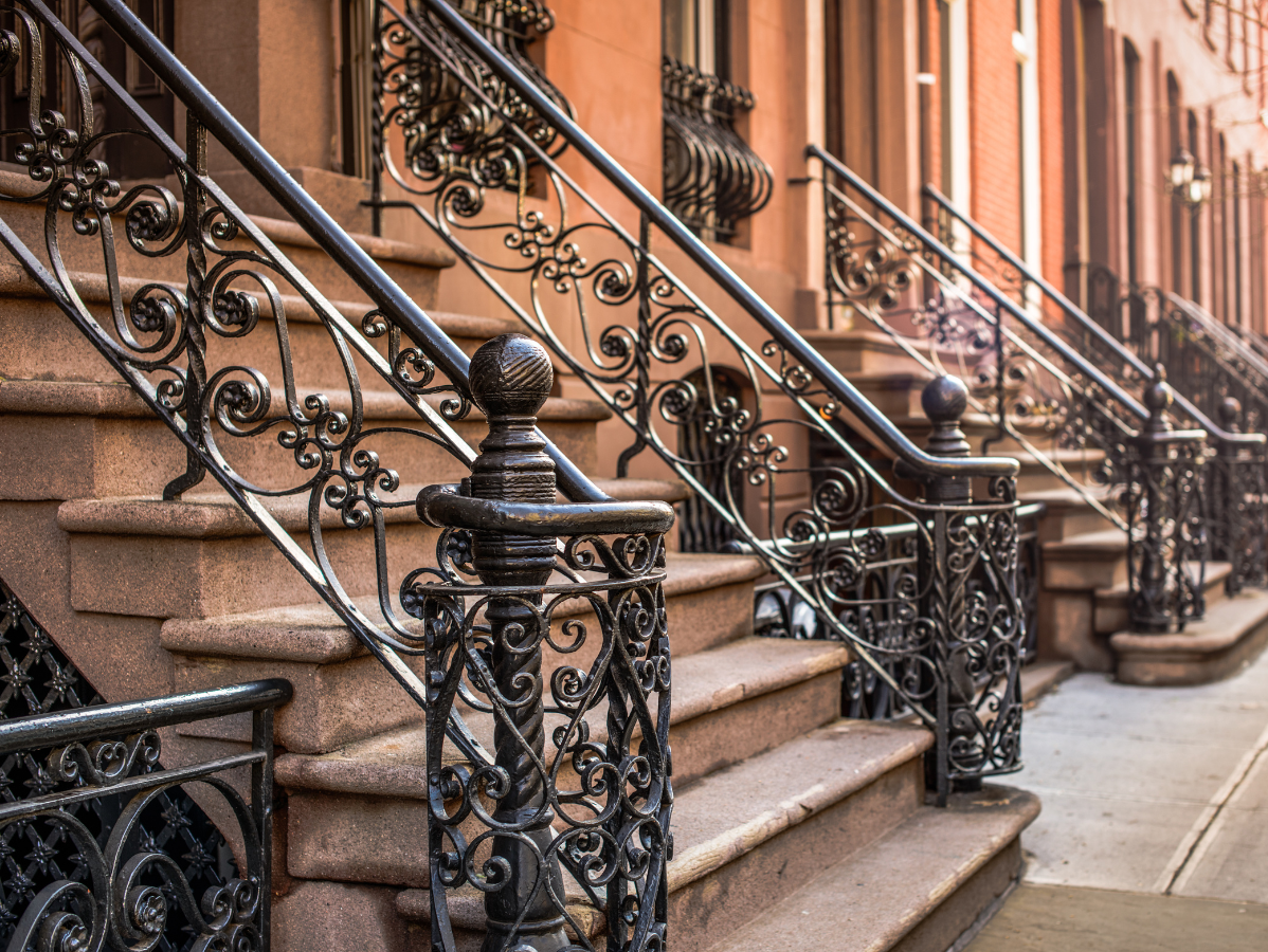 Brownstone steps with ornate wrought-iron railings lead to building entrances in a city.