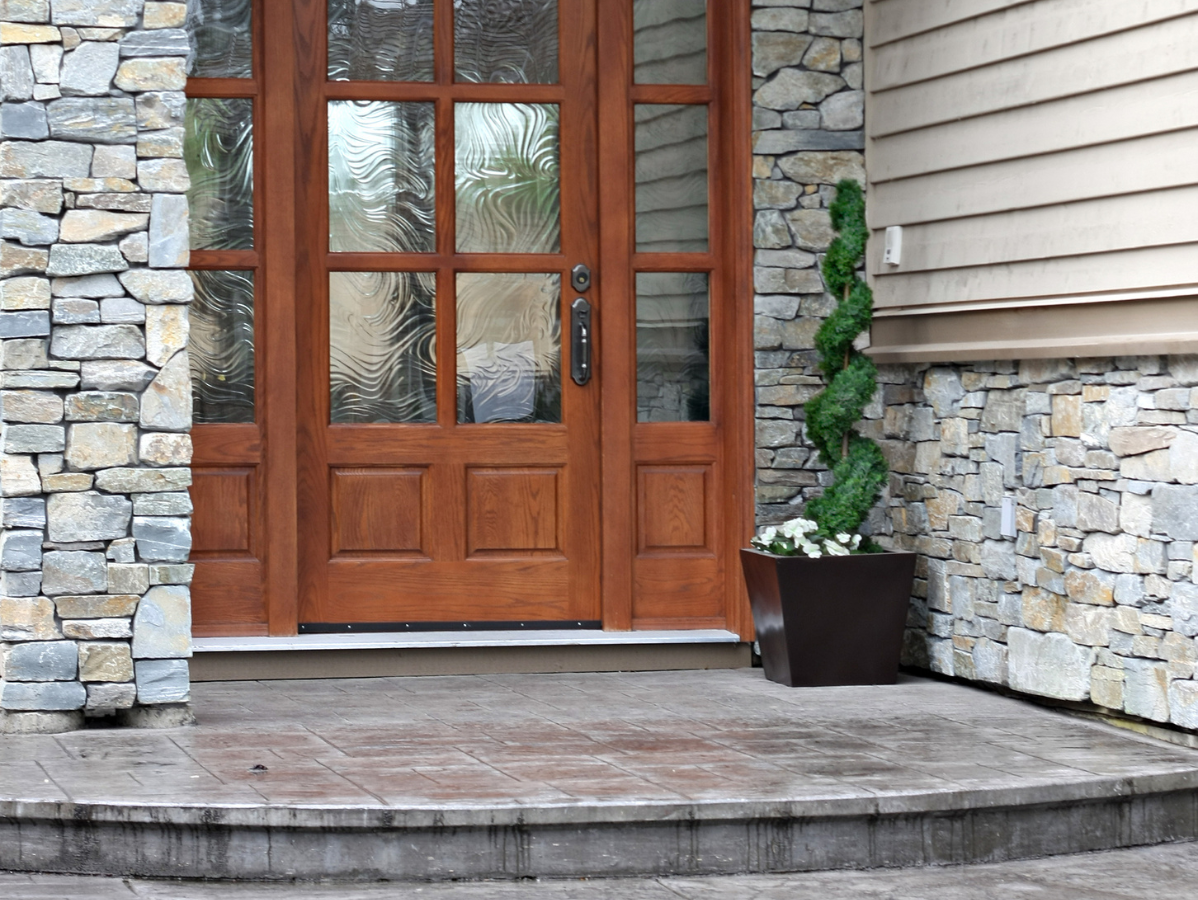 Wooden front door with sidelights, stone facade, and spiral topiary in a planter.