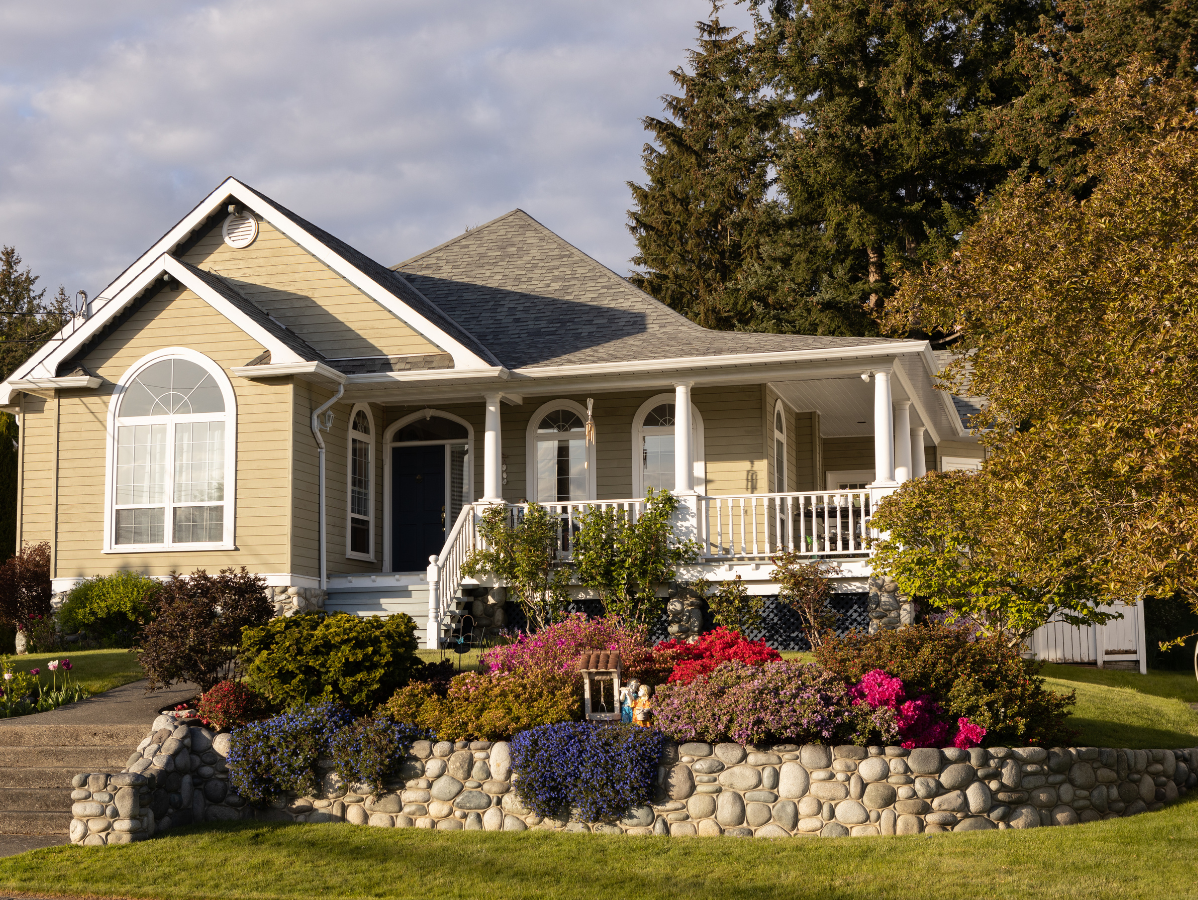 Tan house with porch and colorful garden, stone wall, and trees.