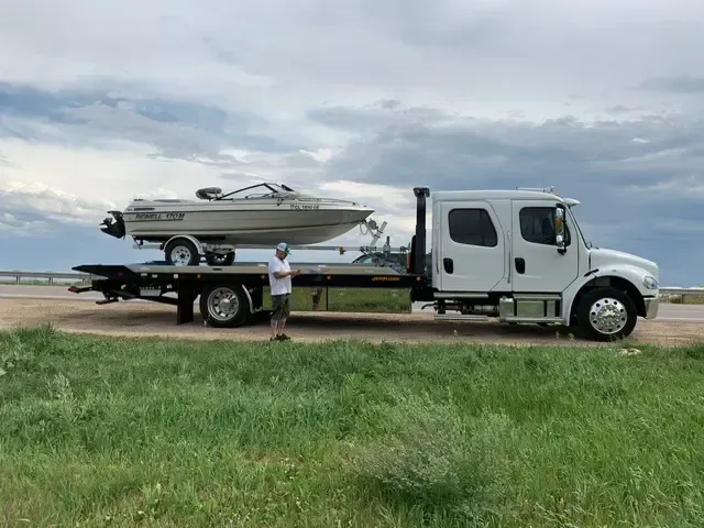 A tow truck carrying a boat on a trailer, man standing beside it, outdoors.