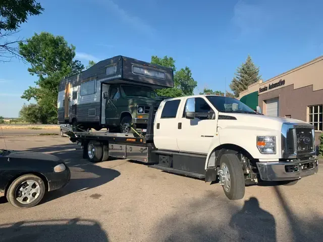 A white tow truck carrying a green RV on its flatbed. Parked next to a gray building.