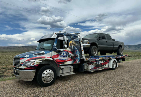 Tow truck with American flag design carrying a gray pickup truck on a rural road.