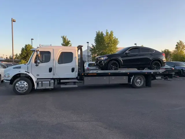 White tow truck carrying a black BMW SUV on its flatbed, parked outdoors.