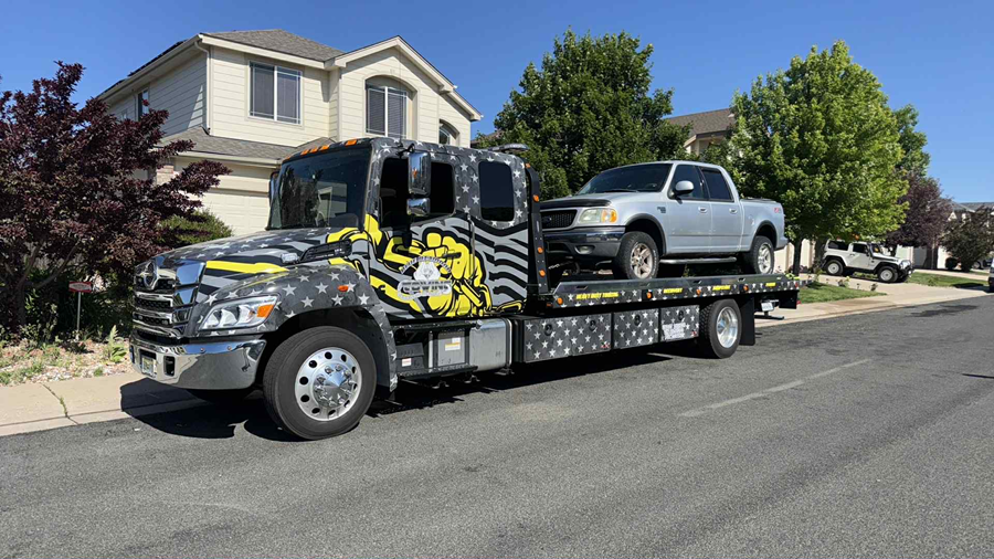 Tow truck with a silver pickup truck on its flatbed, parked on a residential street.