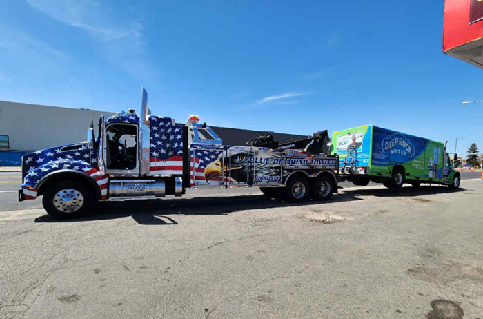 American-themed tow truck towing a green box truck under a blue sky.