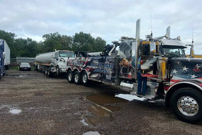 A decorated tow truck towing a white tanker truck on a gravel lot under an overcast sky.