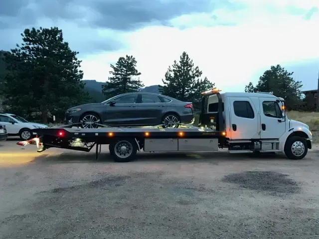 A gray car on a flatbed tow truck at dusk. Truck is white. Other car visible. Mountainous background.