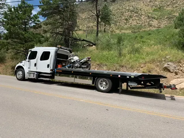 White tow truck with motorcycle on flatbed, parked on a road beside a grassy hillside.