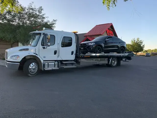 White tow truck carrying a black car in front of a building with a red roof.